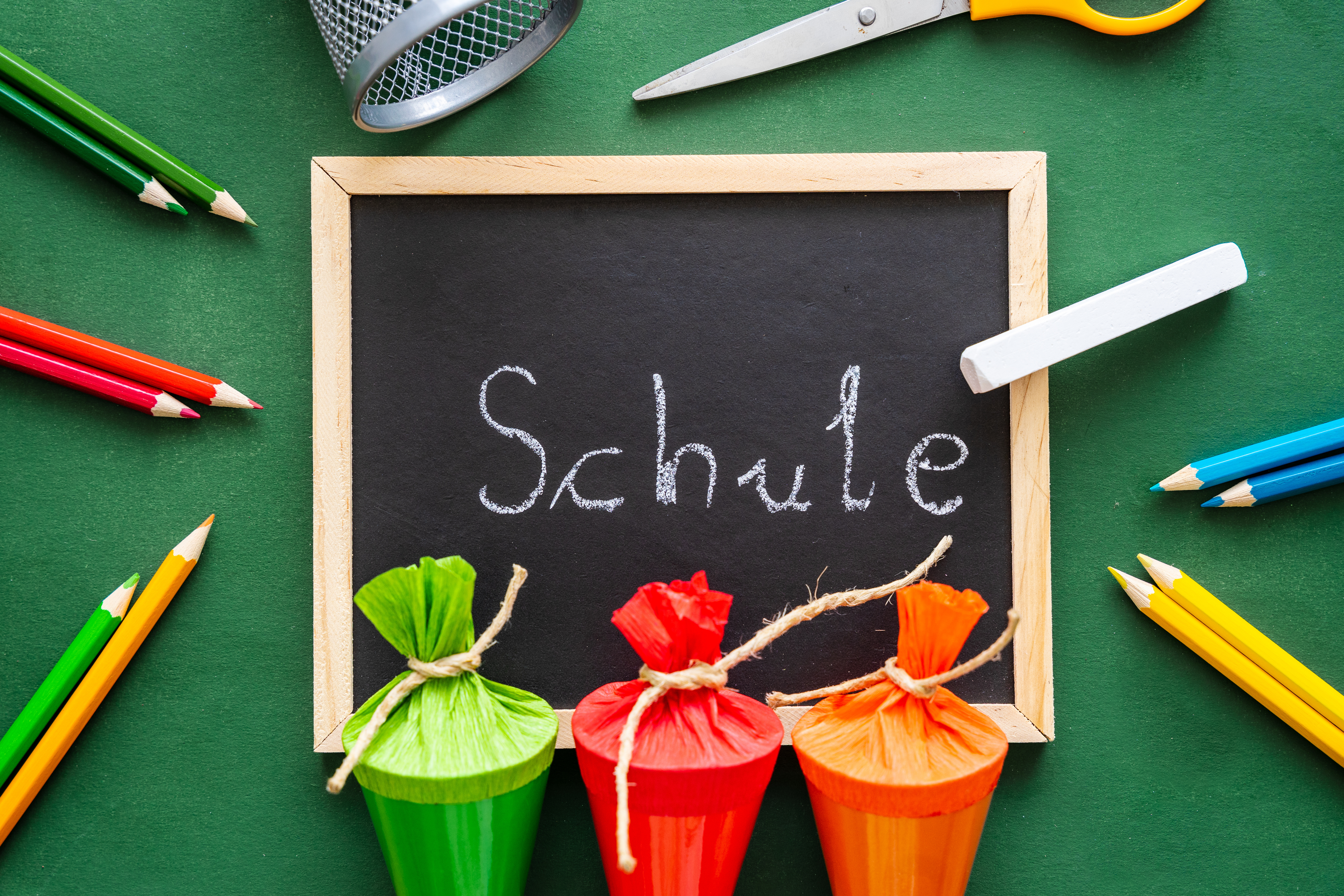 Eine Kreidetafel mit Holzrahmen auf welcher das Wort Schule steht. Um die Tafel herum befinden sich Buntstifte und Schültüten in verschiedenen Farben. 