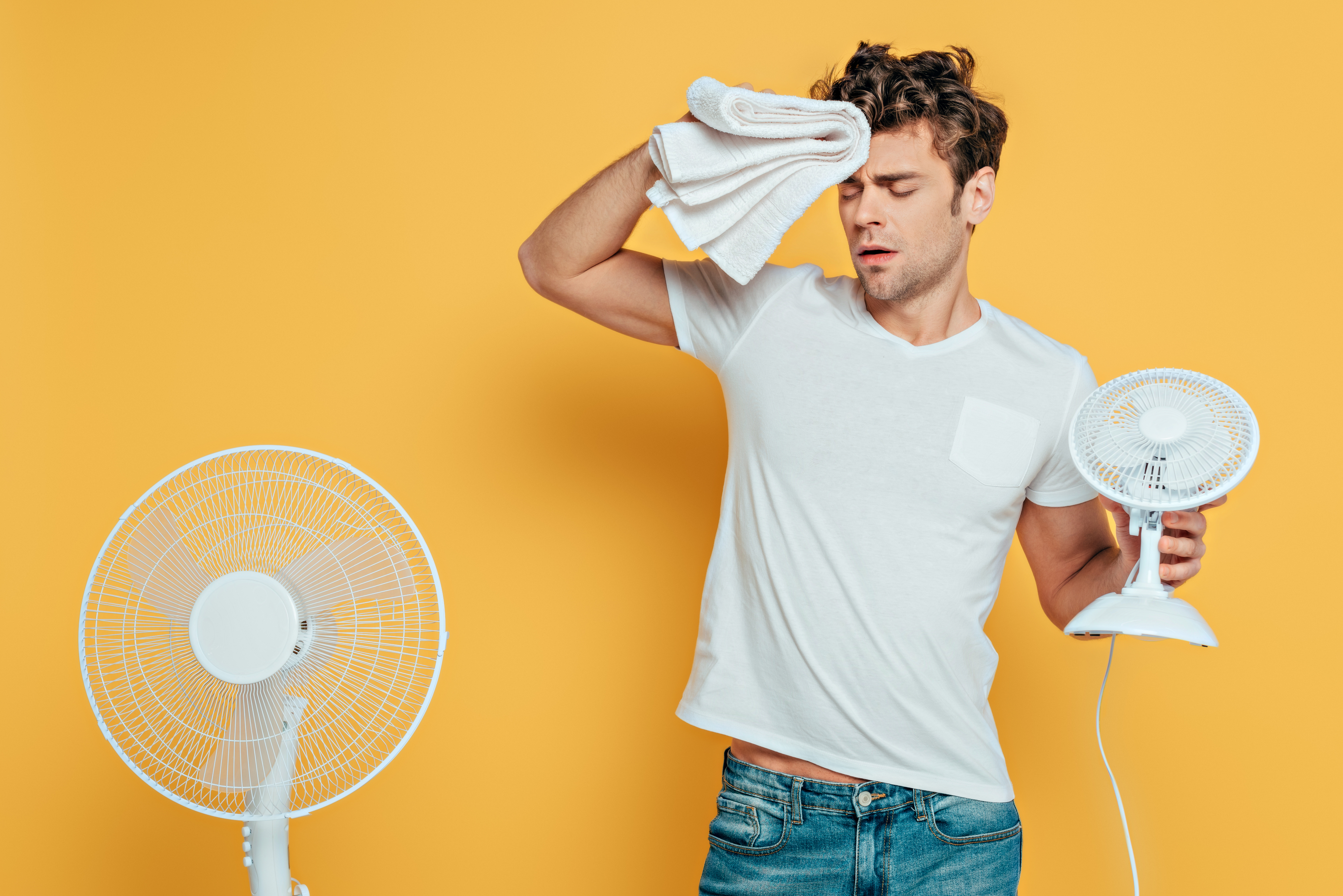 Ein Mann mit Handtuch und Ventilatoren, dem augenscheinlich sehr warm ist und der schwitzt, vor gelbem Hintergrund.